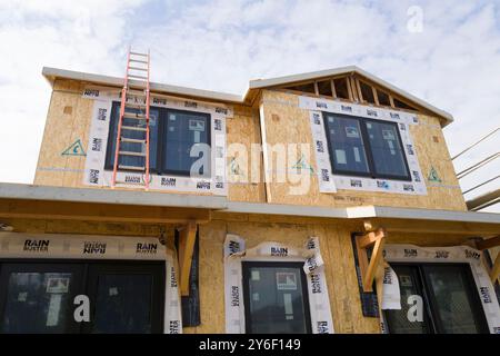 New homes under construction with scaffolding and plywood walls. Workers on-site, building in progress, surrounded by trees and construction equipment Stock Photo