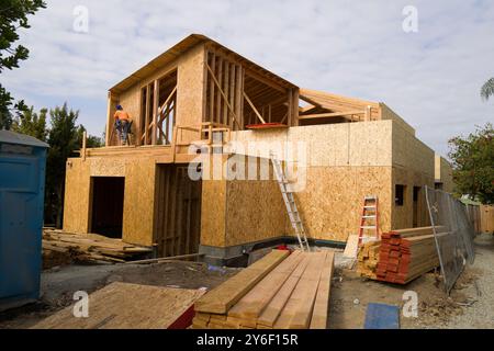 New homes under construction with scaffolding and plywood walls. Workers on-site, building in progress, surrounded by trees and construction equipment Stock Photo
