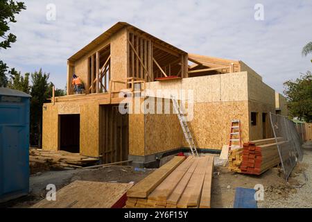 New homes under construction with scaffolding and plywood walls. Workers on-site, building in progress, surrounded by trees and construction equipment Stock Photo