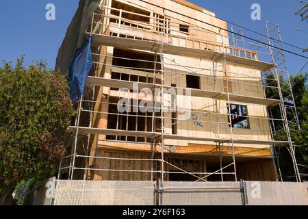 New homes under construction with scaffolding and plywood walls. Workers on-site, building in progress, surrounded by trees and construction equipment Stock Photo