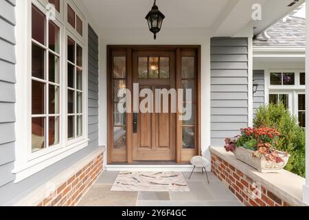 A front door and covered porch detail on a home with grey siding, white trim, red brick, a wooden front door. Stock Photo