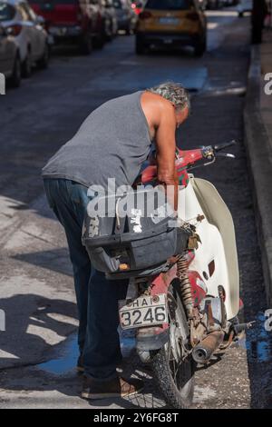 older man preparing or mending small motorcycle at the roadside, elderly gentleman fixing his scooter or moped on a street side in Zante town, Greece. Stock Photo
