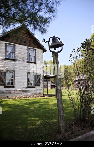 A crooked old antique cast iron dinner bell is mounted on a wooden post outside a house in an American Pioneer village. Stock Photo
