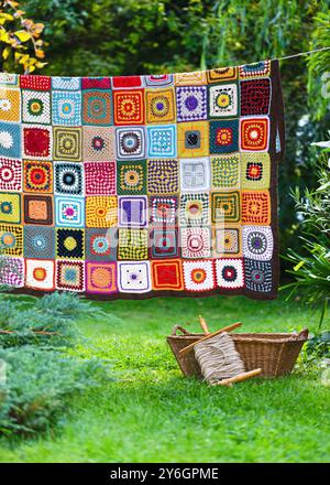 Colorful handmade crochet blanket made from woolen granny squares hanging outdoors on the string with rustic laundry wicker basket. Stock Photo