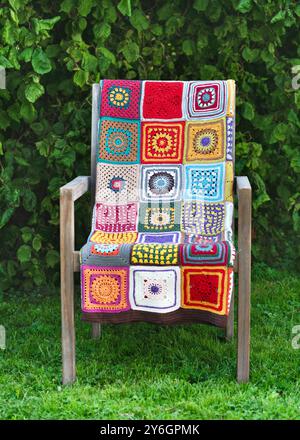 Bright beautiful multi colored plaid, make from crochet granny squares with different motifs, lies on a wooden chair in the cottage garden. Copy space Stock Photo