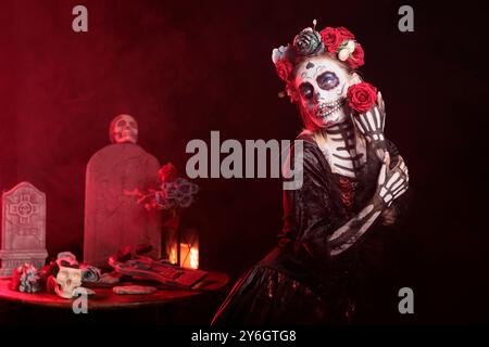 Graceful woman at Day of the Dead event with spooky decorations to remember the deceased. Lady holding red rose for traditional holiday of remembrance for those who have passed, red light background Stock Photo
