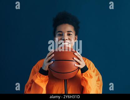 Portrait of a young female with curly hair holding a basketball and smiling against a blue backdrop Stock Photo