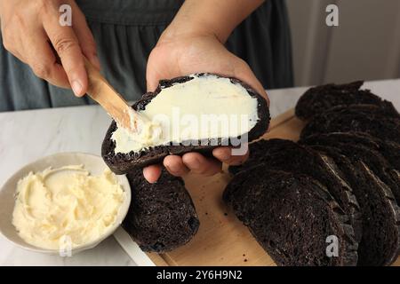 A Woman Spreads Cream Cheese with a Wooden Cutlery Knife to the Chocolate Sourdough Bread.  Woman Hands Spreading Cream Cheese on Bread Slice. Stock Photo