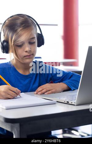 In school, boy wearing headphones writing in notebook and using laptop Stock Photo