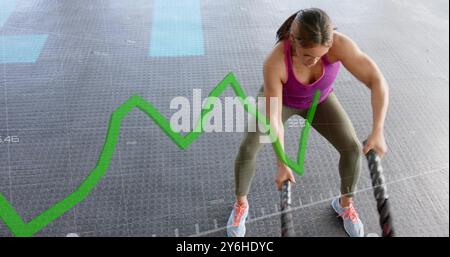 Green line graph image over woman exercising with battle ropes in gym Stock Photo