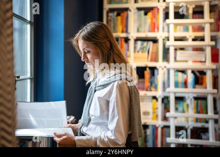Interested focused middle aged woman student reads book standing near window in university library. Stock Photo