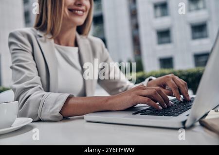 Pleasant working day. Close up of young woman using laptop and smiling while working indoors Stock Photo