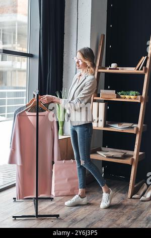 Successful fashion stylist. Full length of attractive young woman in smart casual wear choosing clothes from the rank and smiling while standing in th Stock Photo