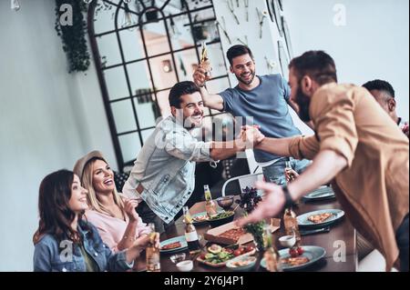 Among best friends. Handsome young man toasting and smiling while two men shaking hands Stock Photo