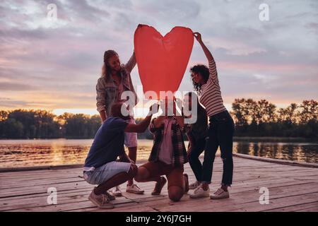 Heart shaped lantern. Full length of young people in casual wear lighting up sky lantern while standing on the pier Stock Photo