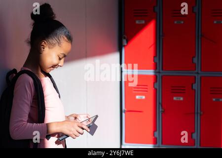 Using smartphone, girl standing by lockers in school hallway, smiling Stock Photo