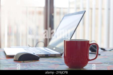 A red coffee mug sits on a table next to a laptop computer. The laptop is open and the screen is blank. The scene suggests a quiet and focused atmosph Stock Photo