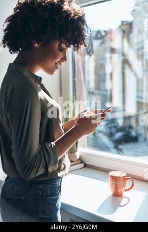 Enjoying nice chat with friend. Attractive young African woman using her smart phone and smiling while standing near the window indoors Stock Photo