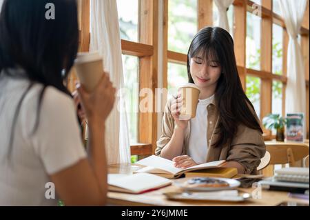 Two cheerful young Asian college students are enjoying coffee while reading together in a minimalist coffee shop. people, lifestyles, university life, Stock Photo