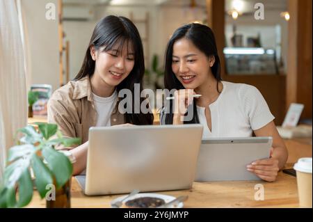 Two positive young Asian female friends are working remotely together from a coffee shop, discussing work while using a laptop and a digital tablet. Stock Photo