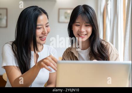 Two positive and charming young Asian female friends are working remotely together from a coffee shop, discussing work while looking at a laptop scree Stock Photo