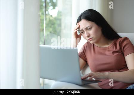 Worried asian woman checking bad news on laptop sitting on a couch at home Stock Photo
