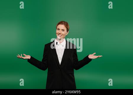 A confident TV presenter in sleek black speaks enthusiastically to the audience with a vivid green backdrop. Stock Photo