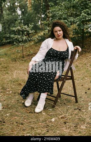 A plus size woman with curly hair relaxes on a chair in a serene field adorned with greenery. Stock Photo