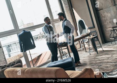 Nice to meet you. Two young fashionable men shaking hands and smiling while standing in workshop Stock Photo