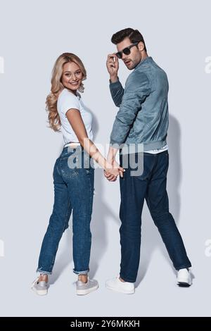 Having fun. Full length of beautiful young couple looking over shoulder and smiling while standing against grey background Stock Photo