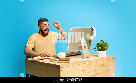 Young emotional bearded man sitting at desk and interacting with computer, expressing excitement against blue studio background Stock Photo