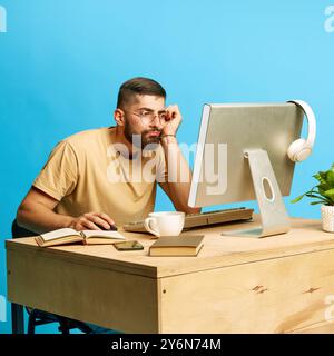 Young bearded man sitting at desk and looking on laptop, scrolling social media and online services against blue studio background Stock Photo