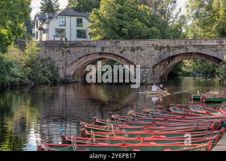 A stone bridge spans a river and is reflected in the water. Rowing boats for hire are moored to the riverbank and a couple row on the river. People ob Stock Photo