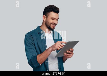 Technologies in everyday life. Charming young man using his digital tablet and smiling while standing against grey background Stock Photo