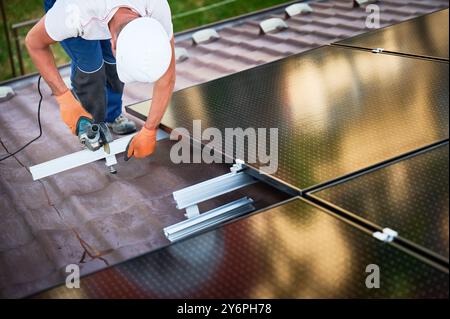 Worker installing or maintaining photovoltaic solar panel system on roof of house. Technician in helmet using grinder on metal part of the panel's mounting structure. Concept renewable energy. Stock Photo