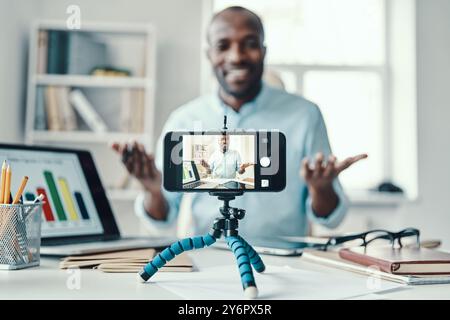 Happy young African man in shirt showing chart and smiling while making social media video Stock Photo
