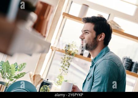Nice morning. Handsome young man in casual wear drinking coffee and smiling while standing in the kitchen at home Stock Photo