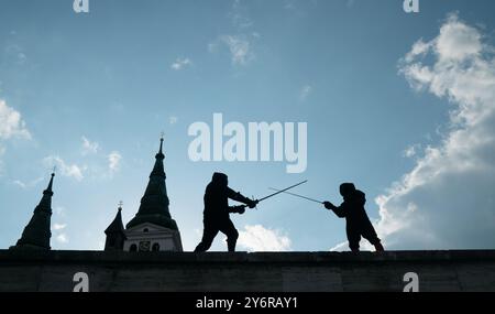 Two fencers dressed in black protective uniforms, helmets with face masks are fencing on castle wall using long medieval metal historical swords pract Stock Photo