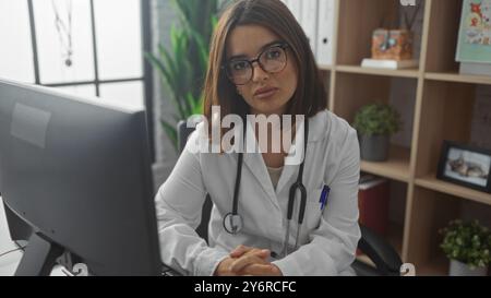 A young, attractive, hispanic, brunette woman in a white coat with a stethoscope, seated at a desk in a clinic workplace, looking at the camera with a Stock Photo