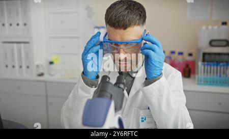 A focused man with a beard wearing safety goggles and gloves uses a microscope in a medical laboratory. Stock Photo