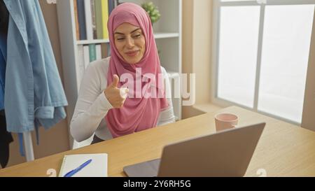 A smiling woman wearing hijab gesturing thumbs-up at a desk with a laptop, in a bright home office setting. Stock Photo