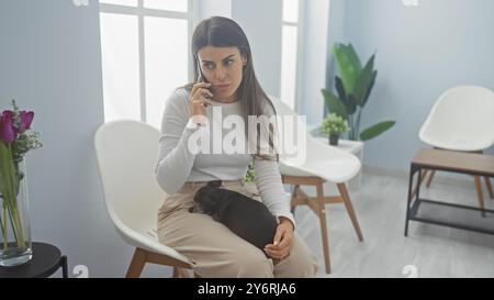 Hispanic woman talks on phone holding dog in a bright vet clinic waiting room Stock Photo
