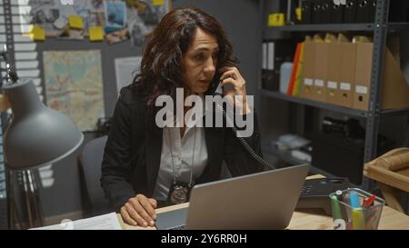 A middle-aged woman detective is working at her desk in an office, talking on the phone and using a laptop Stock Photo