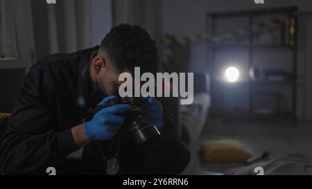 Focused hispanic detective man examines evidence at indoor nighttime crime scene with camera. Stock Photo