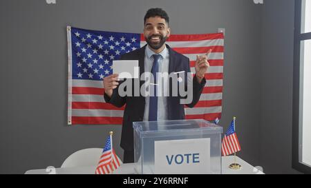 A smiling young hispanic man points while holding a ballot at a us electoral college with an american flag backdrop. Stock Photo