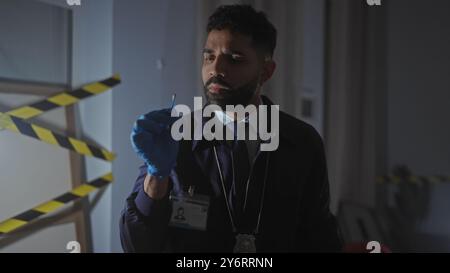 A bearded hispanic detective man examines evidence at a nighttime indoor crime scene with caution tape in the background. Stock Photo