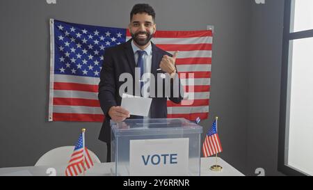 A smiling young hispanic man with a beard giving thumbs up after voting in an american indoor electoral setting with flags. Stock Photo