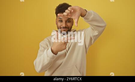 A cheerful man makes a camera frame gesture against a bright yellow wall, evoking creativity and positivity. Stock Photo