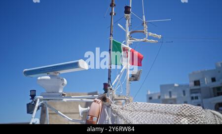 Italian flag waves on a boat against a clear blue sky in monopoli, italy, with maritime radar and white buildings in the background. Stock Photo