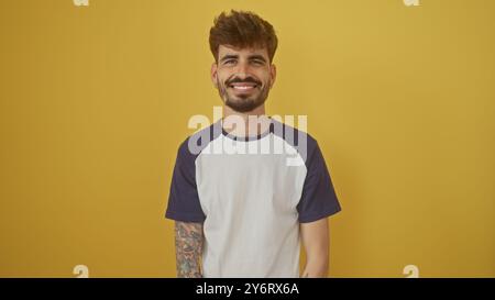 Handsome young man with beard and tattoos smiling confidently against a vibrant yellow background Stock Photo